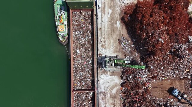 A large boat is at the dock unloading metal scrap. A crane is moving materials next to the water. The area is filled with various debris and is clearly part of an industrial operation.