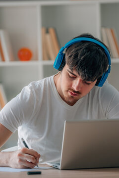 student with laptop and headphones at home studying online and writing or taking notes