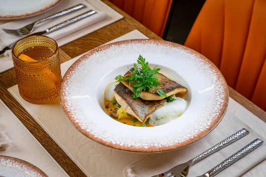 Pan seared white fish fillet served in restaurant table setting. Wide angled shot of seared fish fillets with vegetables and foam, alongside a glass and cutlery on a table.