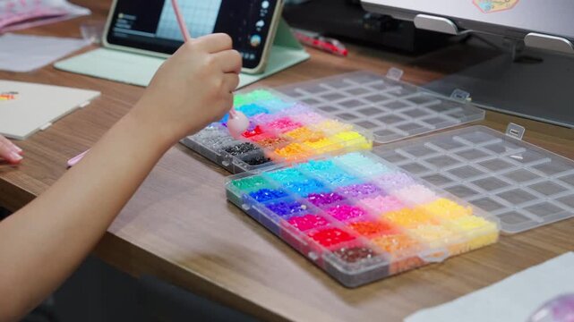 A wide shot of a young girl focused on assembling colorful plastic fuse beads on her workspace table, with an iPad displaying reference designs in the blurred background.