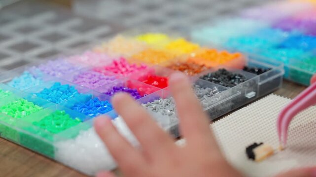 Foreground shot focused on a multicolored bead box, with a young girl's hands actively using tweezers in the blurred bokeh background.