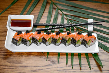 Salmon sushi rolls topped with black caviar on white plate. Salmon maki sushi rolls with black roe on white rectangular plate, green palm leaf background, studio lighting. © Анна Терелюк
