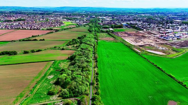 Aerial view of vibrant green farmland and a tree line in the West Coast of Scotland, with a typical town expanding into the rural landscape