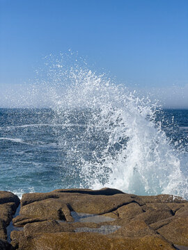 A spectacular close-up of a sea wave exploding into a high white splash against coastal rocks under a clear blue sky. Dynamic nature concept.