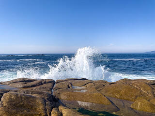 Fototapeta premium A dynamic seascape capturing a powerful sea wave exploding into white foam and high splash against brown coastal rocks on a sunny day. Great for travel and nature themes.