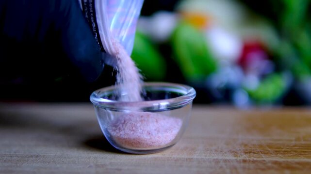 Pouring pink Himalayan salt into glass bowl with fresh vegetables on background healthy cooking ingredient