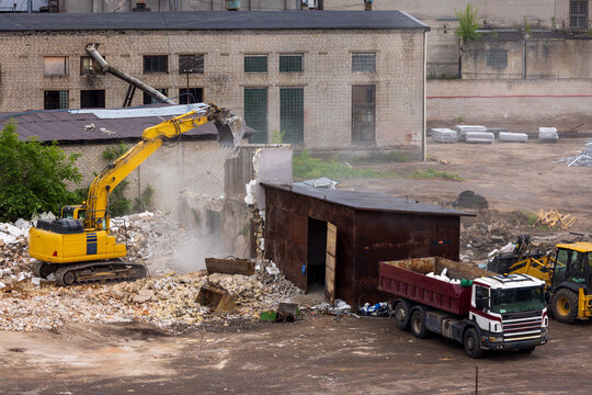 View of a yellow excavator demolishing a brick structure next to a red dump truck at a construction site with rubble and dust Vilnius, Vilnius County, Lithuania.