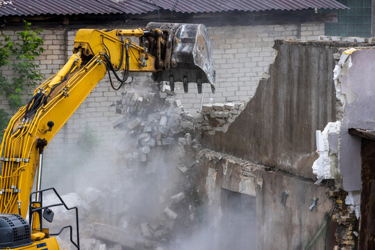 View of a yellow excavator demolishing an old brick wall with dust and debris falling during construction work in Vilnius, Vilnius County, Lithuania.