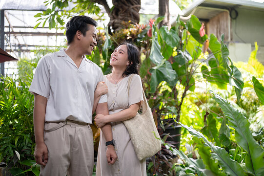 Happy asian man and woman couple linking arms while walking through in green tropical garden at cafe