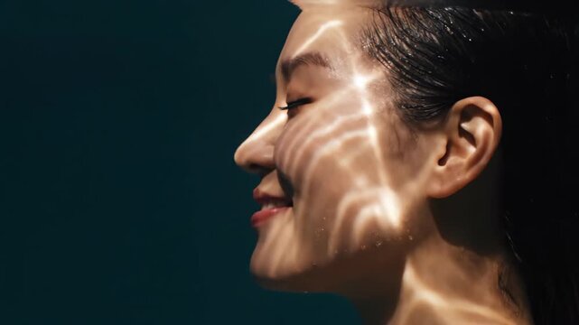 Asian woman submerged in water with dynamic light refraction on skin. Side view close-up of face enjoying water therapy. Skin care and wellness concept in motion