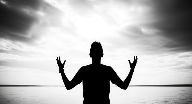 Silhouette of a man with arms outstretched facing a dramatic cloudy sky over the ocean black and white
