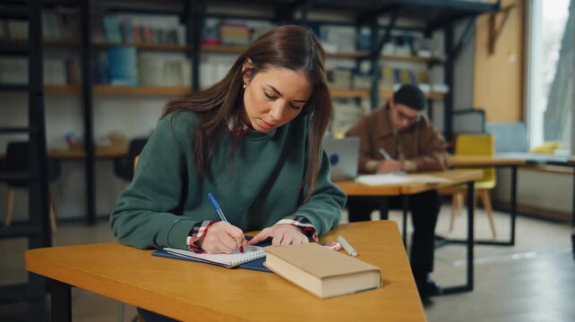 Students study in a library with books and workstations during the day, focusing on their tasks quietly in a shared learning space