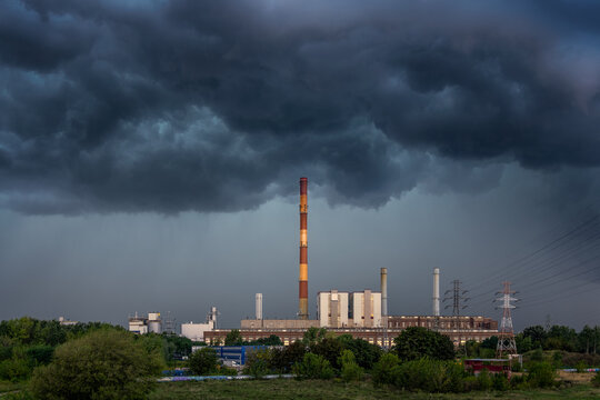 View of a coal power plant with a tall red and white striped chimney and industrial buildings under a dramatic dark stormy sky in Warsaw, Masovian Voivodeship, Poland.
