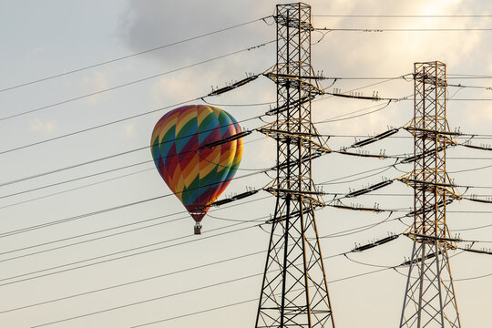 View of a colorful rainbow hot air balloon floating behind high-voltage power lines and transmission towers against a clear sky in Warsaw, Masovian Voivodeship, Poland.