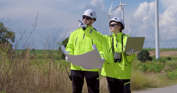 Two Caucasian engineer in bright safety jacket discussing blueprint near wind turbine farm under clear sky analyzing project data outdoor, renewable energy teamwork innovation clean power generation