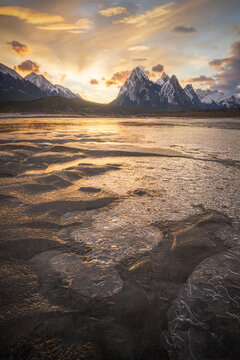 View of the frozen surface of Abraham Lake with snow-capped mountains under a golden sunset sky in Clearwater County, Alberta, Canada.