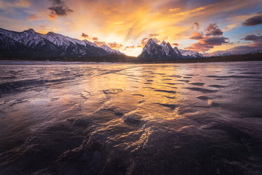 View of the frozen surface of Abraham Lake with snow-capped mountains under a vibrant sunset sky in Clearwater County, Alberta, Canada.