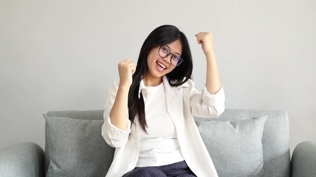 Happy and cheerful young Asian woman celebrating her success or achievement, dancing with raised clenched fists while sitting on the sofa in the living room at home.