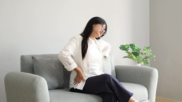 Young Asian woman sitting on the sofa in the living room of her house, massaging her back feeling pain, sore, stiff, cramped, stretching her body, tired.