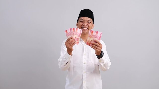 Asian Muslim man wearing a peci and white shirt, carrying and showing a wad of Indonesian rupiah banknotes, happy to get profit, salary, THR concept.