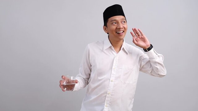 An Asian Muslim man wearing a peci and white shirt, standing while carrying a glass of water, looking at the clock, waiting for the time to break the fast, hearing adzan.