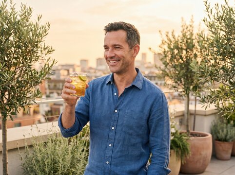 Man smiling and holding a cocktail on a rooftop terrace. Warm sunset city view and relaxed summer atmosphere