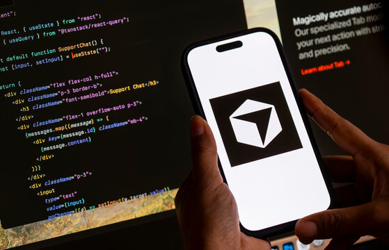 In this photo illustration, The Cursor AI logo is displayed on a smartphone screen in front of computer code and a software development interface