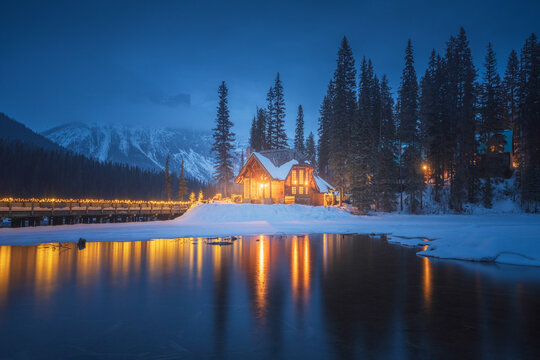 View of a cozy illuminated wooden lodge on a snowy lakeshore with reflections on the water and pine trees under a twilight sky in Jasper, Alberta, Canada.