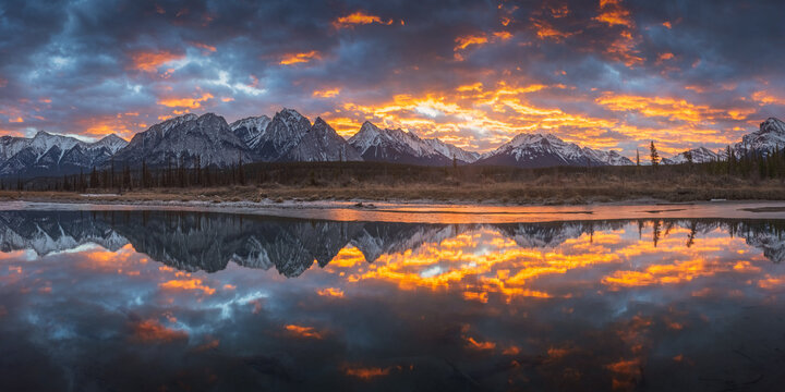 View of snow-capped mountain peaks reflected in the calm water of a river under a dramatic sunset sky with orange clouds along the Icefields Parkway Jasper, Alberta, Canada.