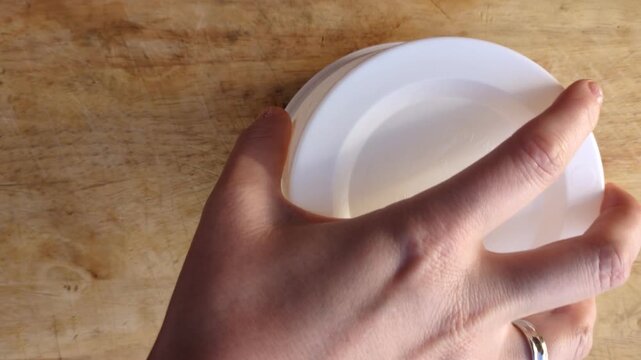 Top view of a person unmolding a wheel of fresh white goat cheese from a plastic mold container onto a rustic wooden board, traditional cheese making and gourmet artisan food production