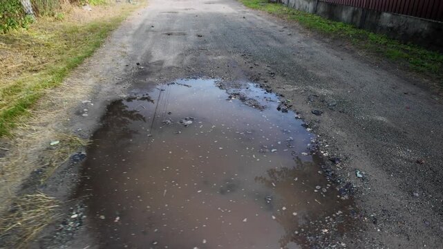 A cinematic 4K 24fps handheld shot of a muddy water puddle on a rough dirt road. Dynamic camera movement creates a realistic motion blur effect, capturing a gritty, realistic rural atmosphere.