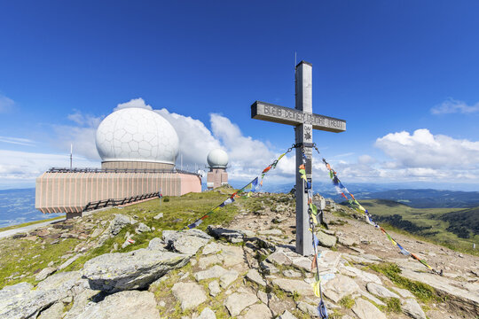 View of the Koralpe mountain summit featuring a large white radar dome and a wooden summit cross with prayer flags under a blue sky in Wolfsberg, Carinthia, Austria.