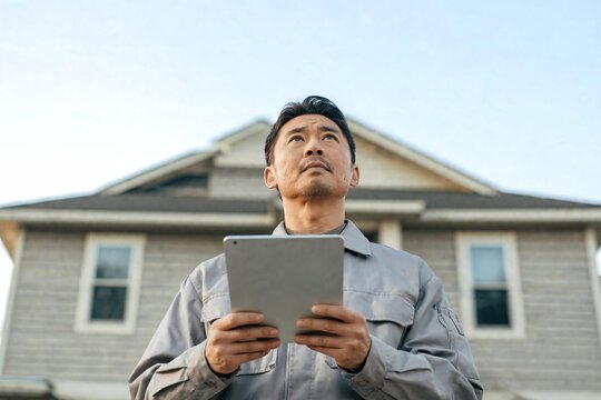 Asian man contractor using a digital tablet for home inspection, surveying a house structure, assessing property condition for renovation and real estate investment