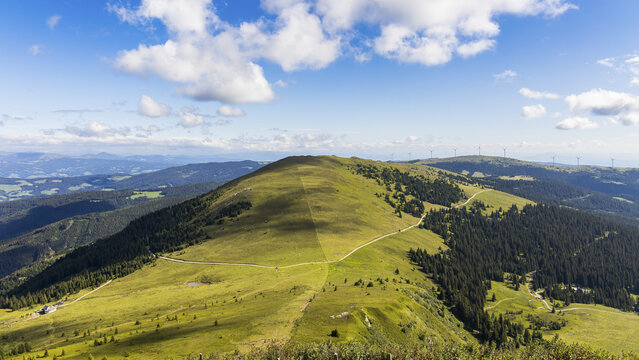 View of the Koralpe mountain range with rolling green slopes, winding dirt paths, and wind turbines under a blue sky in Wolfsberg, Carinthia, Austria.