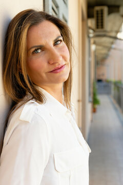 Woman in white shirt leaning against wall in an outdoor corridor, softly smiling. Milan, Italy
