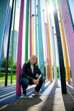 Woman crouching among tall colorful poles with city buildings in the background on a sunny day.