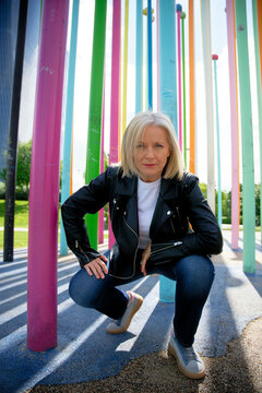 Blonde woman in leather jacket crouching among colorful vertical poles outdoors.