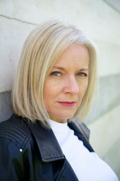 Woman with short blonde hair in a leather jacket leaning against a light-colored wall