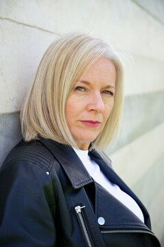 Blonde woman in black leather jacket leaning against a white geometric wall.