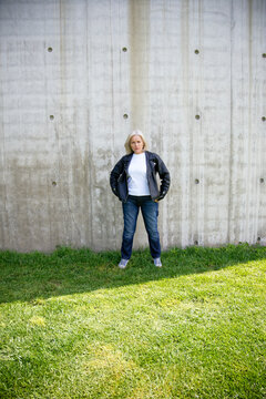 Woman in leather jacket stands confidently against a concrete wall on a grassy lawn.