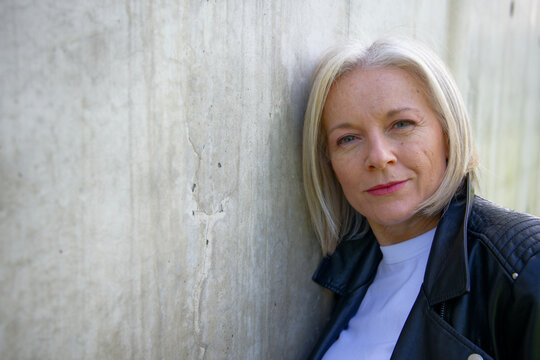 Mature woman with short blonde hair leaning against a textured concrete wall.