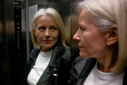 Woman with white hair in black jacket looks at reflection in elevator mirror.