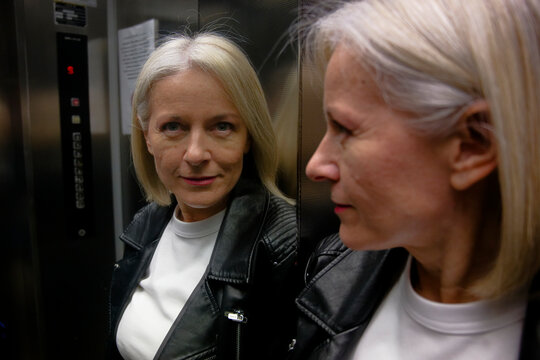 Woman with short blonde hair and leather jacket looks at her reflection in an elevator mirror.
