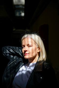 Woman with short blonde hair in black jacket in sunlit interior, looking contemplative.