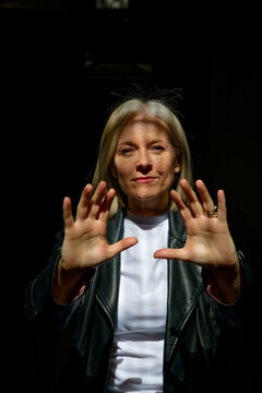 Woman in white shirt and leather jacket raises hands in strong sunlight with a dark background.