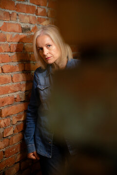 Woman with long blonde hair in denim shirt leans against exposed brick wall.