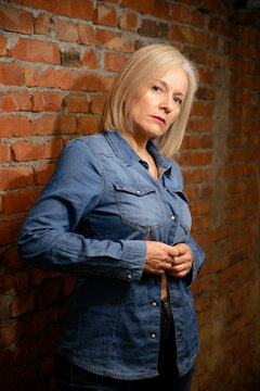 Mature woman in denim shirt standing against a brick wall, looking thoughtful. 