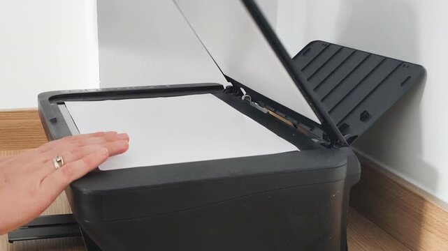 Process of using multifunction printer in corporate office. Human hand closes glass cover of black multifunction printer with white A4 paper inside to scan and copy important business documents.