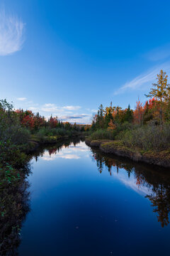 Serene lake reflecting autumnal trees under a clear blue sky with scattered clouds.