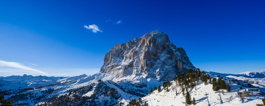 Snow-covered mountain under a clear blue sky surrounded by trees and distant hills.